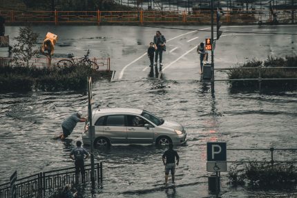 Wide shot of flooded city street, man pushes his car through the water while mom and child watch on despondently